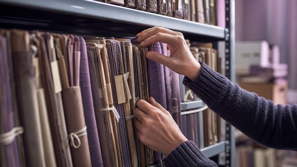 Archivist''s hands selecting a few trusted folders from a crowded records shelf for a clean working tray